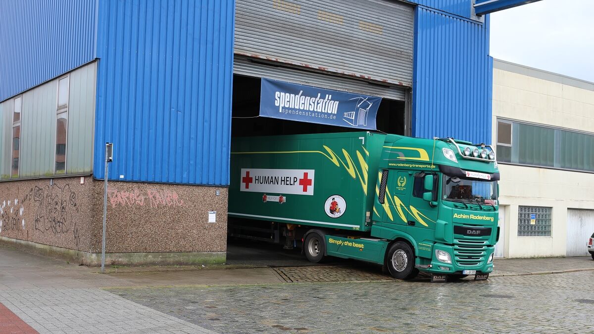 Ein großer grüner LKW von der Spedition Achim Rodenberg aus Celle parkt in der Halleneinfahrt der 12 Meter hohen Bremer Spendenstation. Zu einem Viertel ragt der LKW auf die Straße und auf der Seite des LKW befinden sich Aufkleber mit roten Kreuzen und der Aufschrift Human Help